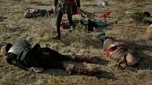 This shows a dry field where soldiers lie on the ground after a battle. A warrior walks among the bodies, surveying the area for any signs of life. The setting is dusty and barren under a clear sky.