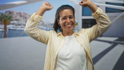 Fototapeta premium Woman hispanic middle aged brunette raising arms and smiling beside yachts at a marina promenade with palm tree and modern building; joyful celebration.
