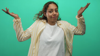 Naklejka premium Woman shrugging with palms up and raised shoulders, wearing yellow striped shirt and white tee in green studio; uncertainty.