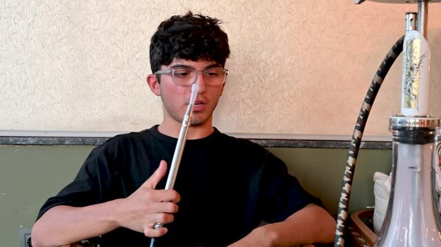 Izmir,turkey,august 10, 2025. Portrait of a young man with curly hair and glasses smoking a shisha pipe while relaxing and talking in a modern hookah lounge