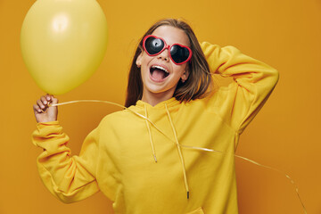 Happy woman in yellow hoodie and red sunglasses holding balloon, smiling with excitement against...