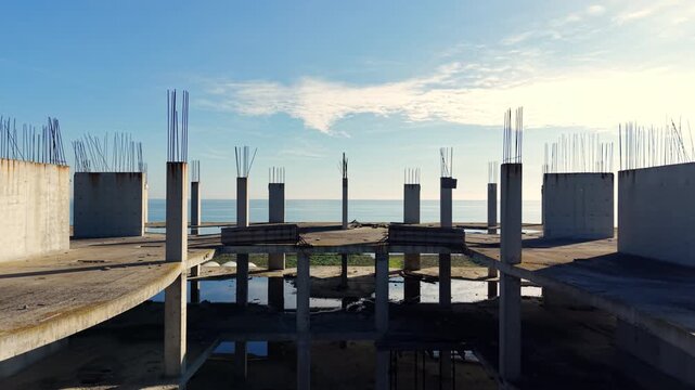 Wide shot of an unfinished building interior showing concrete columns and floor slabs with puddles of water, overlooking the sea at sunrise. Aerial view.