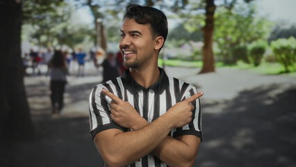Fototapeta premium Young hispanic referee man pointing in opposite directions with fingers in a sunny park, surrounded by green trees and people walking in the background, conveying decision making outdoors.