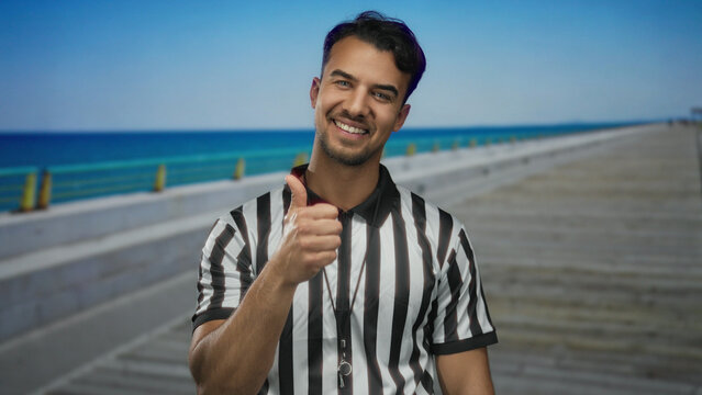 Young hispanic man referee giving thumbs up on promenade near sea outdoors.