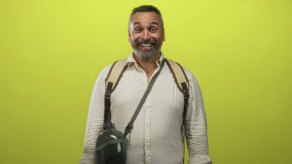 Obraz premium Man middle aged with grey hair and beard with backpack and canteen smiling and laughing in lime green studio; joyful adventure travel.