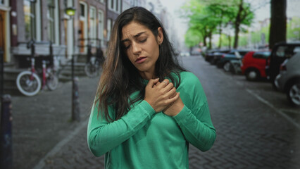 Fototapeta premium Woman clutching chest with both hands on city street near parked cars and bikes in green sweater; distress.