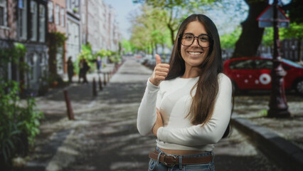 Fototapeta premium Woman giving thumbs up with visible thumb and smiling on a tree lined urban street by a red car and lamppost; optimism approval.