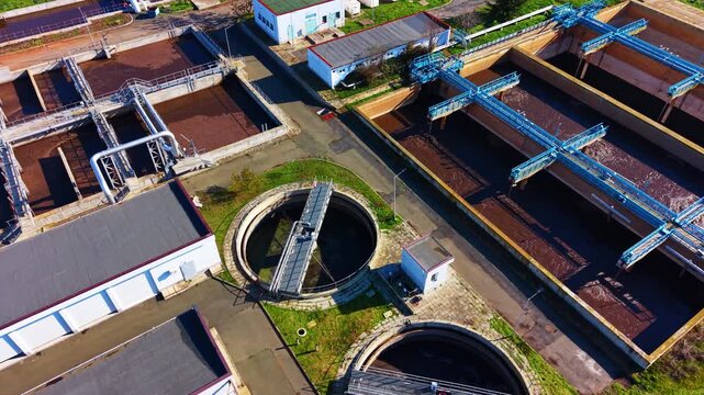 High angle drone shot of sewage treatment facility with rectangular aeration basins, blue metal walkways, and circular clarifier in the foreground.