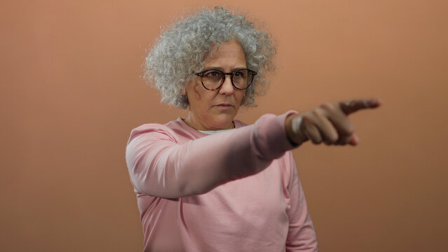 Middle-aged woman with glasses and curly grey hair pointing with focus, wearing a pink sweater against a warm brown background, isolated indoors.
