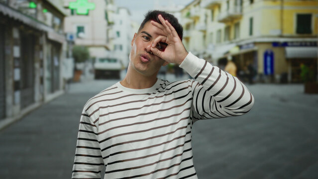 Young man in a striped shirt making an ok gesture in a vibrant city street setting, person exuding confidence and playfulness while standing outdoors during the day.