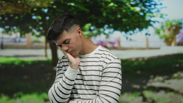 Young man in striped shirt with eyes closed appearing thoughtful in a serene park setting outdoors with greenery and trees in the background suggesting relaxation or daydreaming