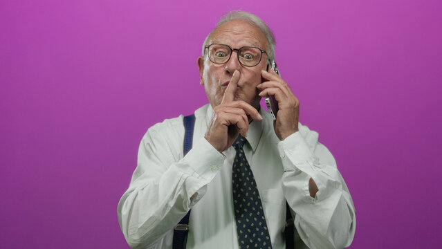 Senior man in business suit gestures silence on phone against pink background, conveying secrecy over call.