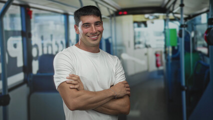 Obraz premium Hispanic man crosses arms inside a color bus interior wearing a white t shirt amid blue seats and metal handrails; denial.