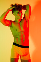 Fototapeta premium Young muscular man posing shirtless in bright colorful yellow and red lighting with hands behind head indoors, curly hair, summer style and toned body emphasize physical fitness and shape.