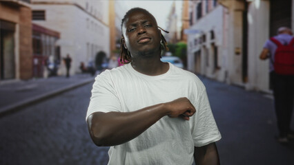 Fototapeta premium Man touching forearm in a white tshirt on a cobblestone street with a pained expression; discomfort concern reflection.