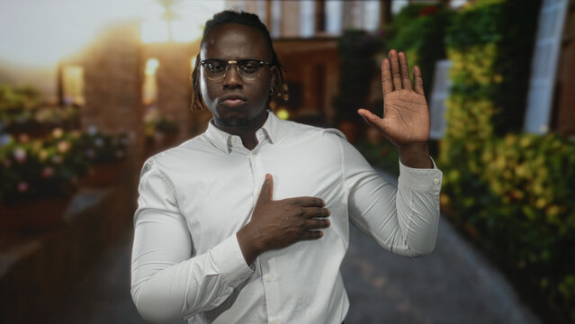 Young african american man with glasses wearing white shirt with hand on chest and raised palm on cobblestone street by brick building and planters; honor pledge respect.