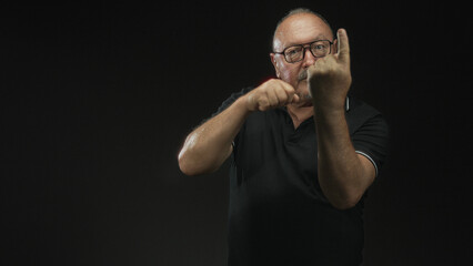 Naklejka premium Man with clenched fists punching air in studio, wearing glasses and a black polo, showing knuckles and forearms against a dark backdrop; determination strength focus.