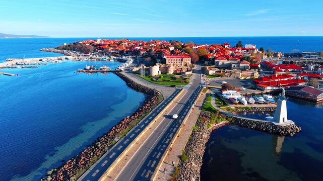 High angle view of the narrow isthmus and road leading to the ancient town of Nessebar, featuring red-roofed houses, a pier, and stone ruins.