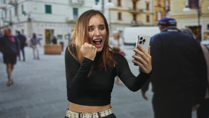Fototapeta premium Young redhead woman holding a smartphone and making a triumphant fist while taking a selfie on a busy street with storefronts and a white van; excitement connection.