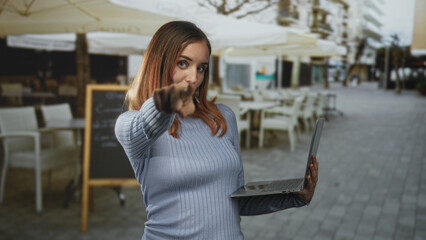 Fototapeta premium Woman pointing finger toward camera while holding laptop on a street cafe terrace beside a chalkboard menu and patio umbrellas; confidence.
