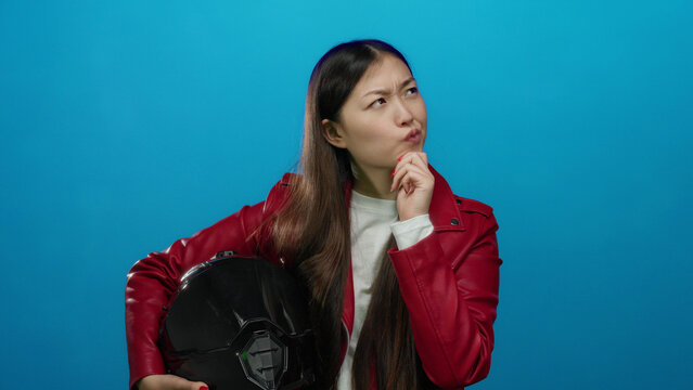 Woman in red jacket pondering holding helmet against blue background with thoughtful expression implying decision-making setting.