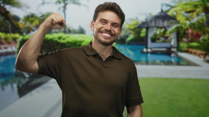 Fototapeta premium Young hispanic man smiling showing teeth and flexing right arm at resort pool outdoors wearing brown polo shirt; joy confidence.