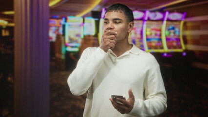Fototapeta premium Man holding smartphone while touching chin in casino with neon slot machines, carpeted aisle and column visible; contemplation decision.