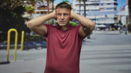 Fototapeta premium Man covering ears with hands at a city street crosswalk near yellow bike racks and buildings; stress sensory overload.