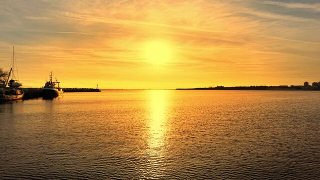 Golden sunset over the sea with fishing boats. Sun rays reflecting on the water surface. Silhouettes of fishing vessels docked at a pier.