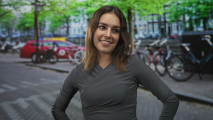 Fototapeta premium Woman smiling with hands on hips on a city street with parked bikes, a red car and trees; confidence warmth curiosity.