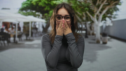Fototapeta premium Woman covers mouth with hands and glasses on urban street near cafe trees, long sleeves visible; surprise reaction.