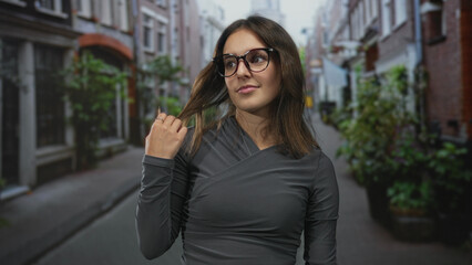 Fototapeta premium Young hispanic woman touches hair while smiling on a narrow cobblestone city street wearing glasses and gray top; confidence calm.