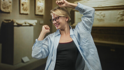 Obraz premium Blond caucasian woman wearing glasses and a light blue shirt raises both fists and dances smiling in a museum building in front of carved stone relief panels; joyful celebration.