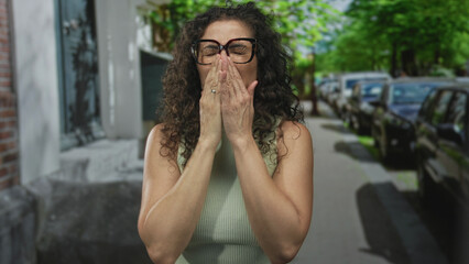 Fototapeta premium Woman clutching chest on street beside parked vehicles and brick building facade under green trees; heartattack.