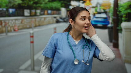 Fototapeta premium Hispanic woman nurse in scrubs with stethoscope listening outdoors on a quiet urban street, capturing a moment of attentive engagement in a contemporary healthcare setting.