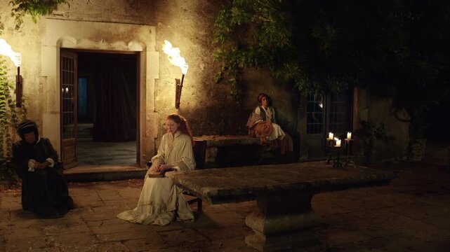 A woman walks through a courtyard lit by torches and candles. Another person sits quietly on a bench. The setting is dark and atmospheric, creating a mysterious mood.