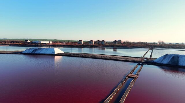 White salt piles situated in pink brine ponds. In the background, green fields and a row of residential buildings under a clear blue sky. Transition between industrial and urban zones.