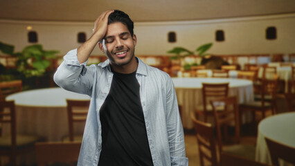 Obraz premium Man with hand on head smiling in a restaurant building with round tables and wooden chairs visible; relief ease.