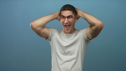 Naklejka premium Man holding hands behind head, mouth open and wearing glasses in a studio with a blue backdrop and light gray t shirt; surprise reaction.
