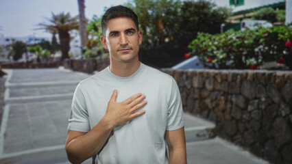 Fototapeta premium Man with hand on chest on street by stone wall and steps in city sunlight, outdoors and standing near plants; sincerity reflection.