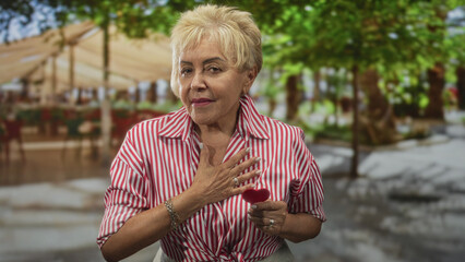 Elderly woman holding small red heart with hand on chest in a street cafe wearing a striped shirt;...