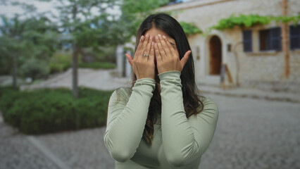 Fototapeta premium Young hispanic woman in olive top covers face with hands smiling in front of stone building; playfulness.