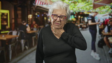 Fototapeta premium Elderly woman holding her throat with one hand and closed eyes at a busy street cafe wearing glasses and black sweater; distress health concern.