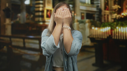 Fototapeta premium Woman blonde covers eyes with hands and makes playful faces while standing in a church building interior near candles and pews; playful surprise.