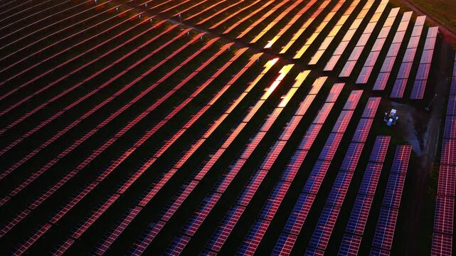 High angle perspective of long rows of solar panels with intense sunset reflections and deep shadows between the rows. Abstract aerial view of solar panels at sunset.