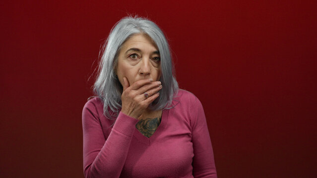 Woman with grey hair covers mouth in surprise against a vivid red background, wearing a pink shirt, conveying emotion and isolation.