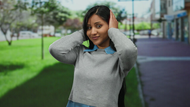 Latin woman in a park covering her ears, wearing a gray sweater and blue shirt, surrounded by lush greenery and urban buildings under a clear sky.