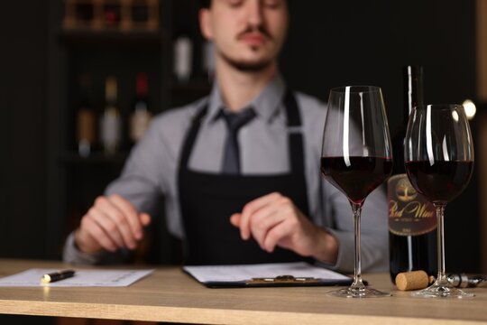 Professional sommelier with wine tasting card and red wine at wooden table indoors, selective focus