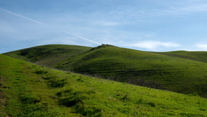 Following the winding, dusty trail as it leads the eye up a steep, vibrant green hillside under a bright, partially cloudy afternoon sky