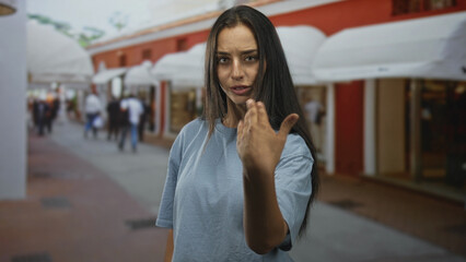 Fototapeta premium Young hispanic woman with hand raised palm out in stop gesture on street lined with storefront awnings; urban defiance.
